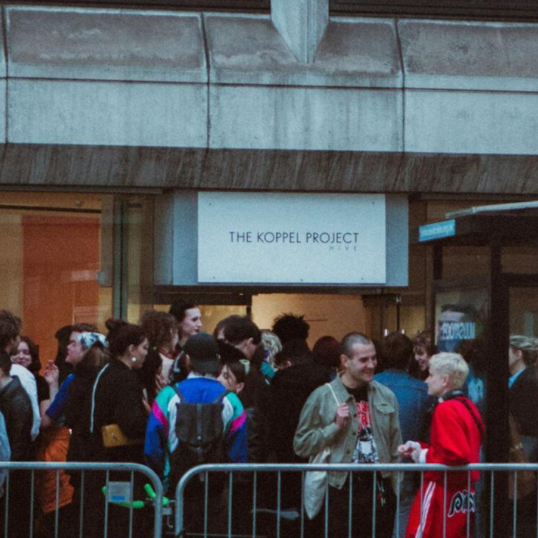 People waiting in line outside a building with 'The Koppel Project' sign.